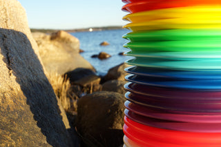 Sixteen disc golf discs in different colors stacked on top of each other with cliffs and water in the background.