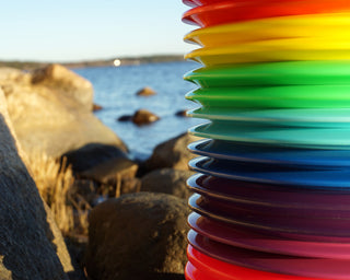 Sixteen disc golf discs in various colors stacked on top of each other with cliffs and water in the background.